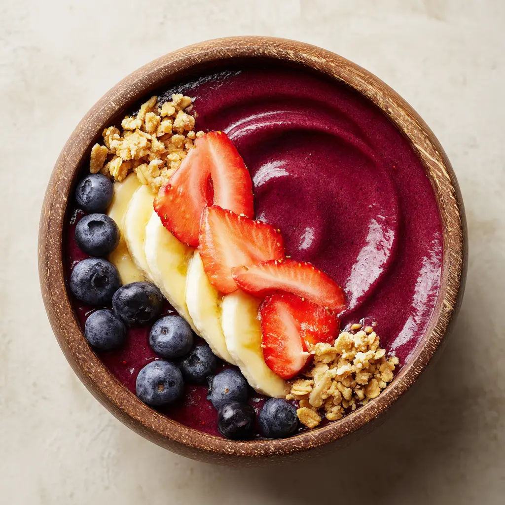 Left rim of an acai bowl topped with a cascade of red strawberries, pale yellow bananas, dark blue blueberries, and golden-brown granola.