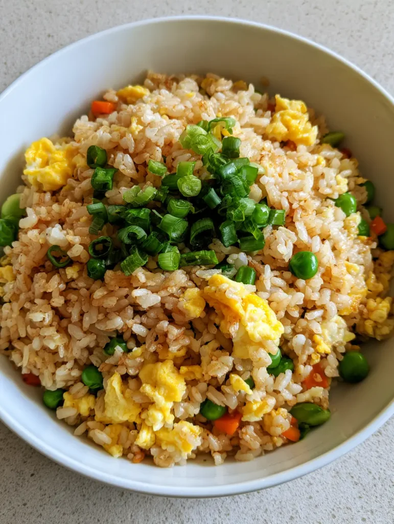 Egg Fried Rice: The Secret to Fluffy, Restaurant-Style Grains at Home 1 Handheld photo of Egg Fried Rice with scrambled egg clusters, sweet green peas, and sliced green scallions on a gray stone surface.