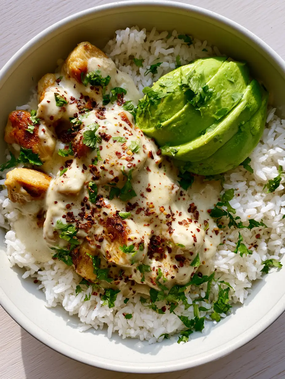 Close up shot of golden-seared chicken breast pieces coated in thick ivory coconut milk sauce inside a white ceramic bowl. (Coconut Chicken Rice Bowl)